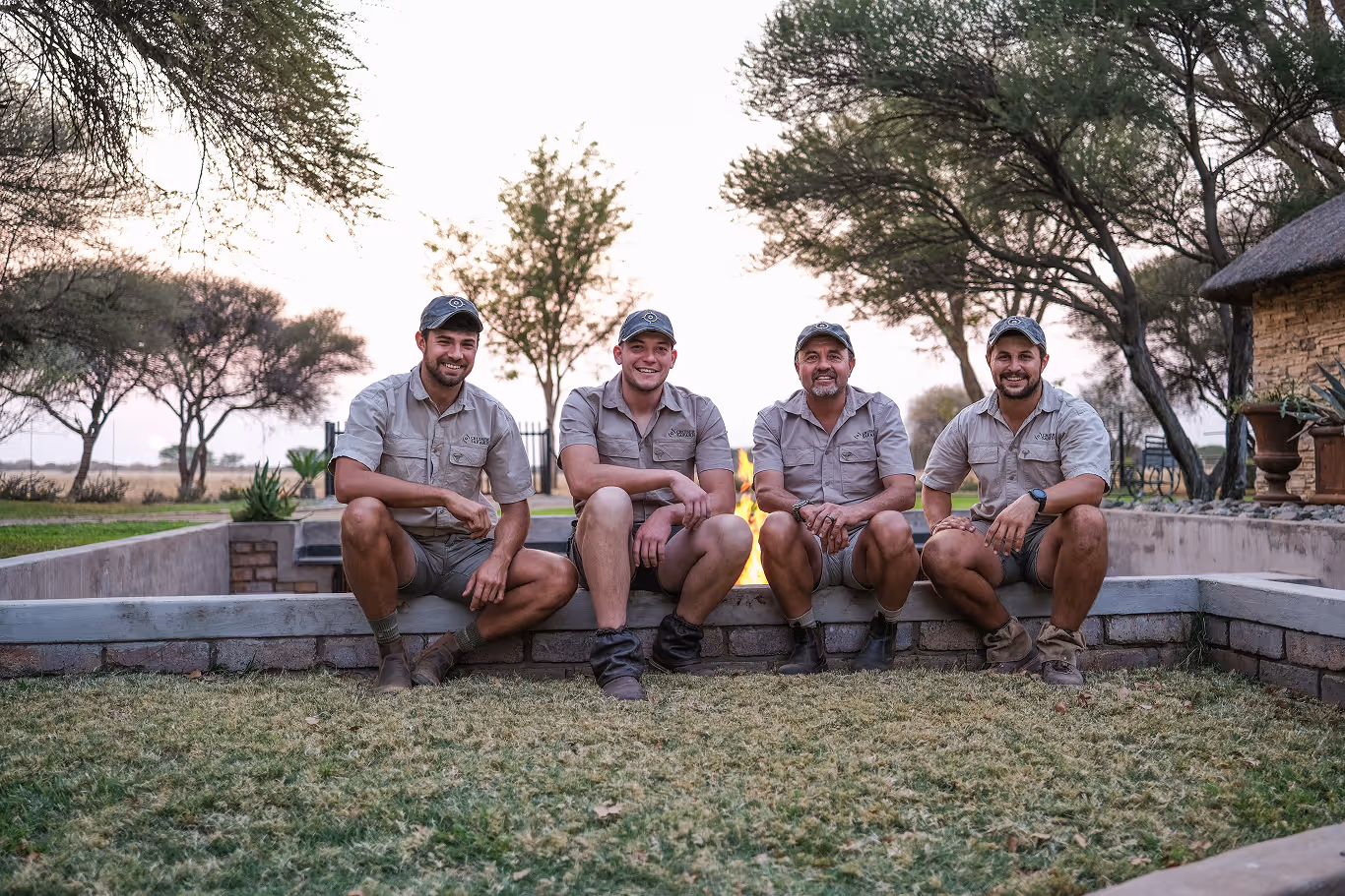 Four men in matching safari uniforms sitting together by an outdoor fire pit